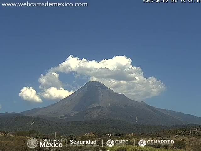 A large, dark-colored volcano is partially obscured by fluffy white clouds under a clear blue sky.