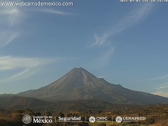 A large, dark-colored volcano is visible under a mostly clear, light-blue sky with a few wispy clouds.