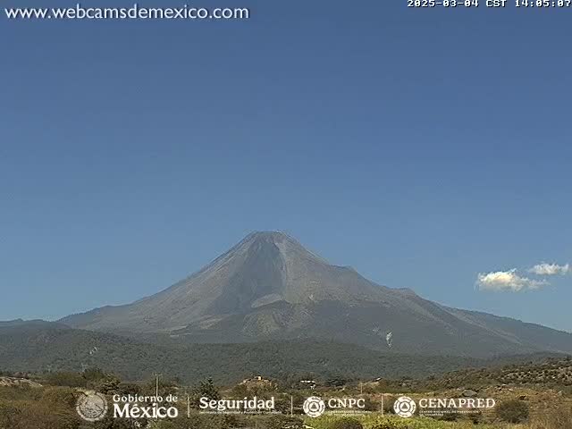 A large, dark-colored volcano dominates the landscape under a clear, bright blue sky with a few small clouds.