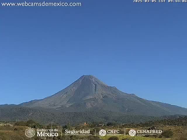 A large, dark-colored volcano is seen on a clear, sunny day, with sparse vegetation at its base.