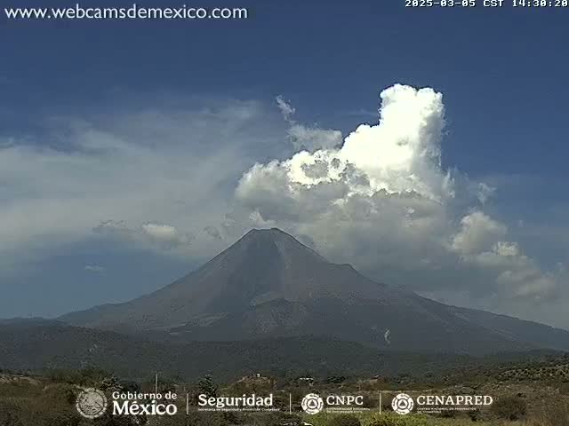 A large, dark-grey volcano is seen under a partly cloudy sky with a prominent cumulus cloud formation.