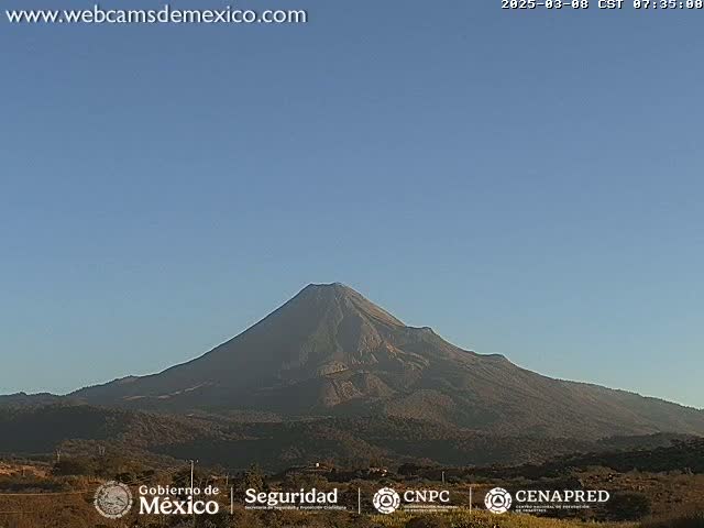 A large, dark-colored volcano is seen under a clear, bright blue sky.