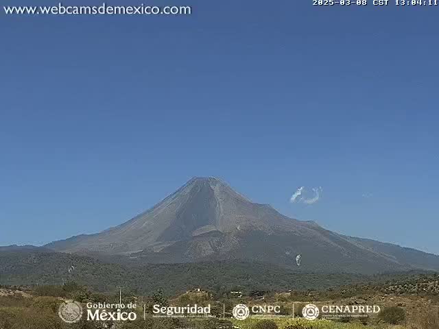 A large, dark-gray volcano is visible under a clear blue sky with a few small, wispy clouds.