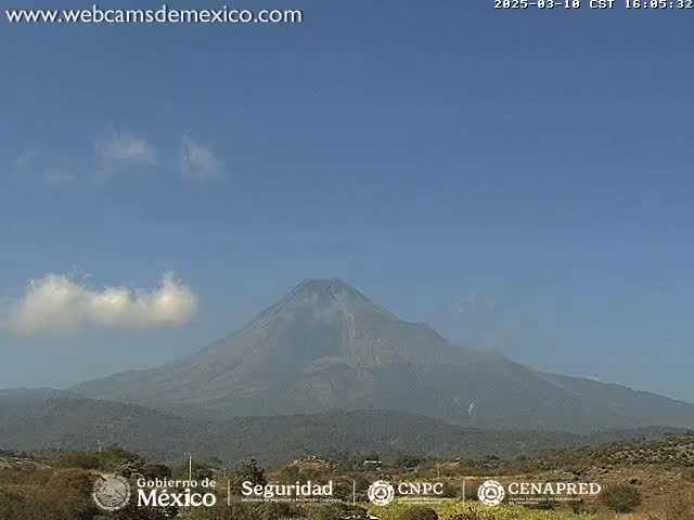 A large, dark-colored volcano is visible under a mostly clear blue sky with a few small, wispy clouds.