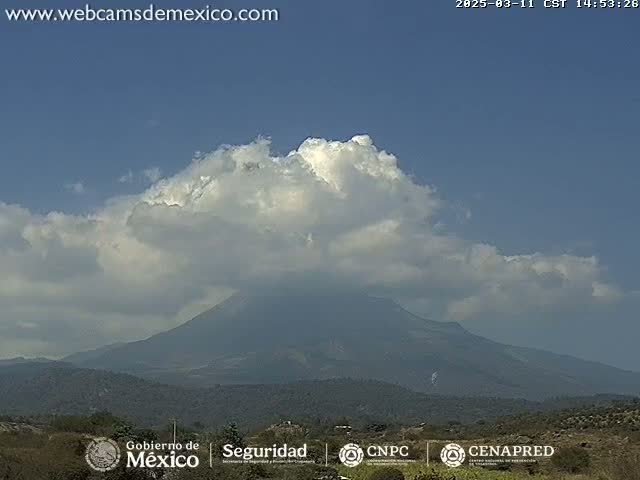 A large, dark mountain is partially obscured by fluffy white clouds under a mostly clear blue sky.