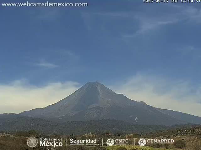 A large, dark-grey volcano is visible under a mostly clear, light-blue sky with a few wispy clouds.