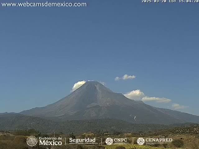 A large, dark volcano is visible under a mostly clear blue sky with a few small clouds.