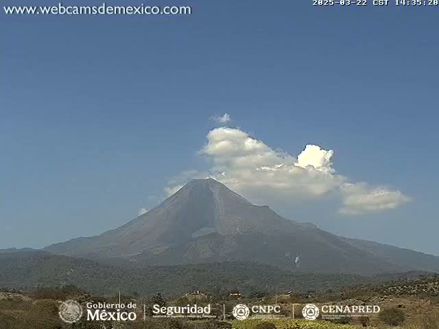 A large, dark volcano is partially obscured by a few fluffy white clouds under a clear blue sky.