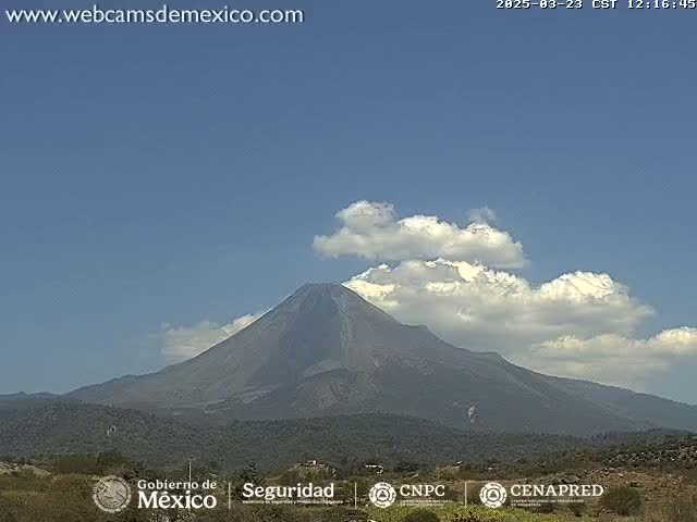 A large, dark volcano is visible under a mostly clear sky with scattered cumulus clouds.