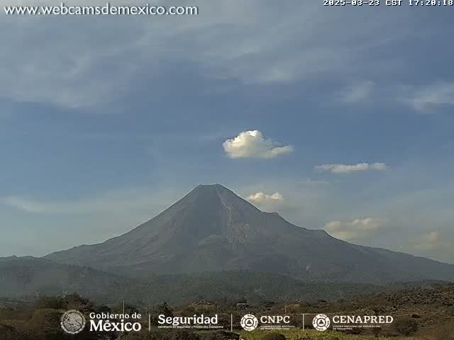 A large, dark-gray volcano is visible under a mostly clear, light-blue sky with a few scattered clouds.