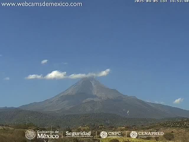 A large, dark-colored volcano with a small plume of smoke is visible under a clear blue sky with a few small, scattered clouds.