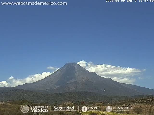 A large, dark-grey volcano is seen under a mostly clear blue sky with some clouds at its peak.
