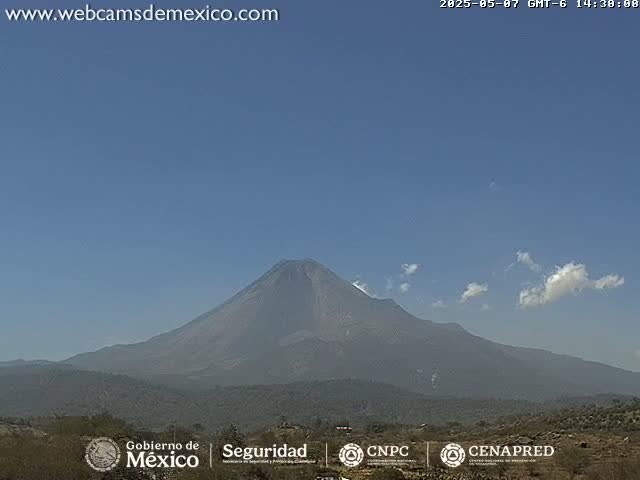 A large, dark-colored volcano is visible under a mostly clear, light-blue sky with a few scattered clouds.