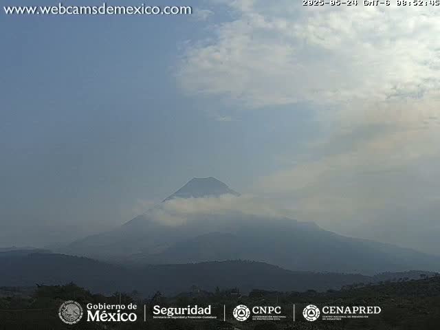 A hazy, partly cloudy day reveals a distant, partially cloud-covered volcano and rolling hills.