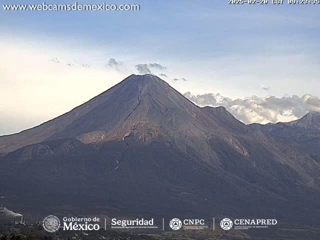 A large, dark-colored volcano with a plume of smoke or steam rises under a mostly clear sky with some clouds.