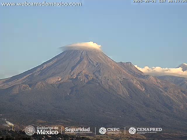 A large, dark gray volcano with a plume of light-colored smoke or cloud emerging from its summit is visible under a clear, light-blue sky.