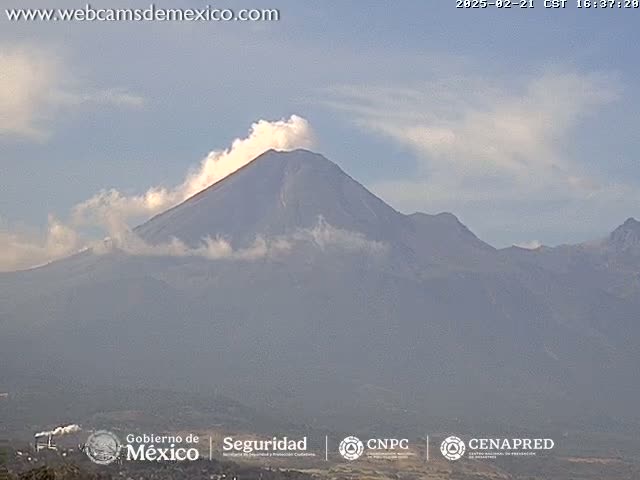 A large, dark-colored volcano is partially obscured by a light-gray haze and a plume of white smoke under a mostly clear sky.