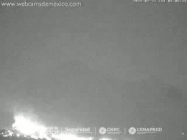 A nighttime, low-light image shows a bright eruption at the base of a dark mountain under a cloudy sky.