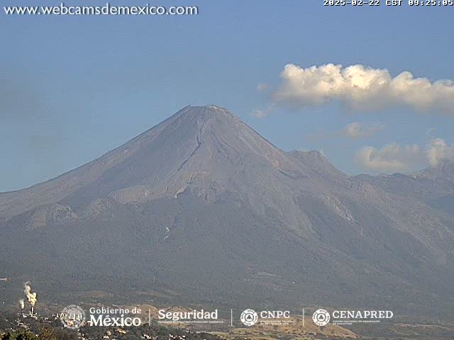 A large, gray volcano is visible under a mostly clear sky with a few small clouds.