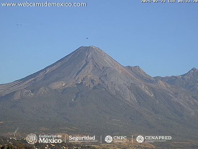 A large, dark gray volcano is seen under a clear blue sky, with a few birds visible in the distance.