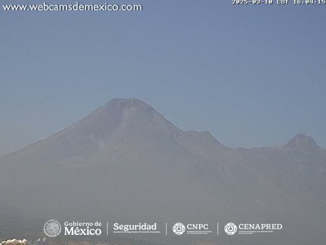 A hazy view of a large, dark-colored volcano under a clear, light-blue sky.