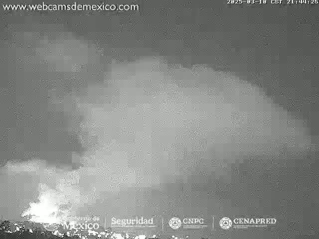 A nighttime, low-light grayscale image shows a volcanic eruption with ash and smoke billowing into the night sky.