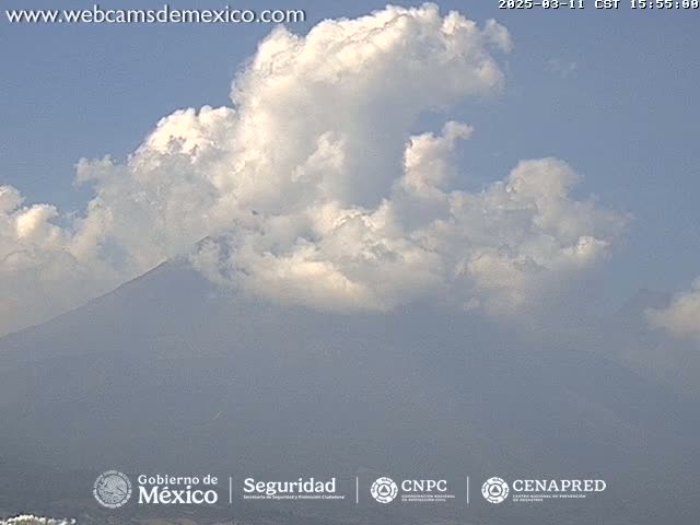 A large, dark mountain is partially obscured by fluffy white clouds under a mostly clear blue sky.