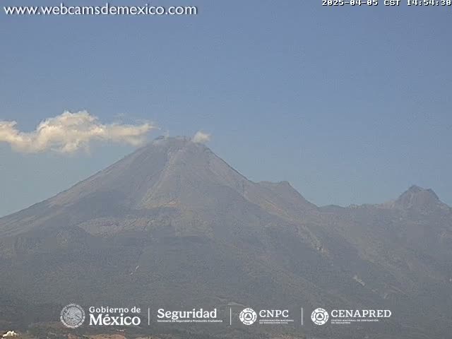 A large, dark gray volcano with a small plume of white cloud near its peak is visible under a clear, light blue sky.