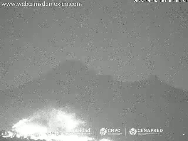 A nighttime, low-light image shows a volcano erupting, emitting a bright, glowing plume of ash and possibly lava against a dark, overcast sky.