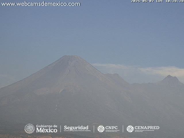 A hazy view shows a large volcano and surrounding mountain range under a clear, light-blue sky.