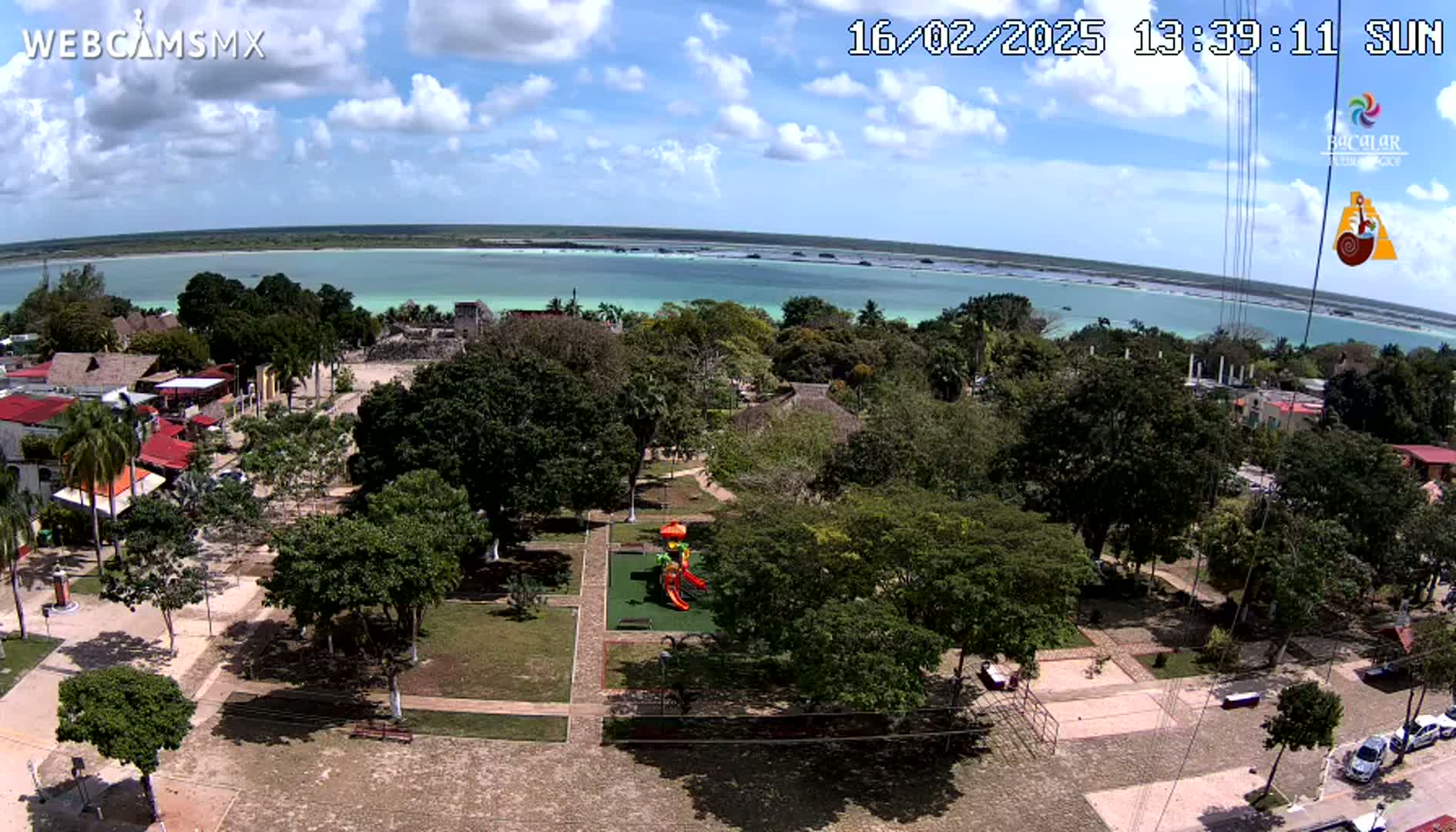 An aerial view shows a town square with lush green trees and a playground next to a calm, light-green body of water under a partly cloudy sky.