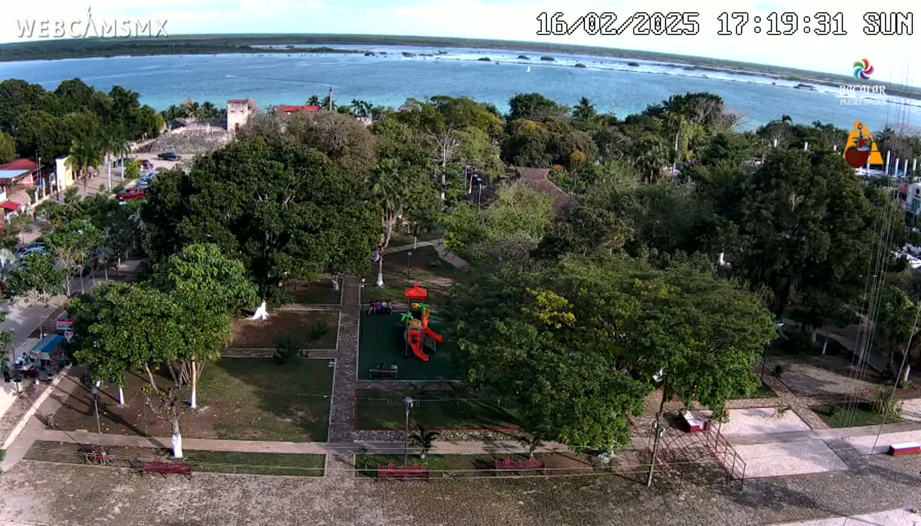 An aerial view of a park with playground equipment, situated near a body of calm water on a sunny day.