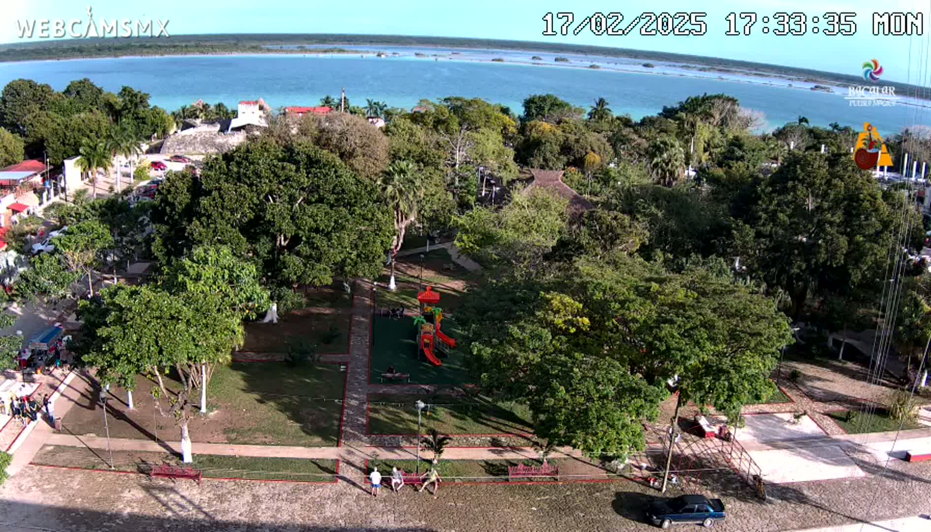 An aerial view shows a park with a playground, surrounded by lush green trees and buildings, overlooking a calm, turquoise body of water under a clear sky.
