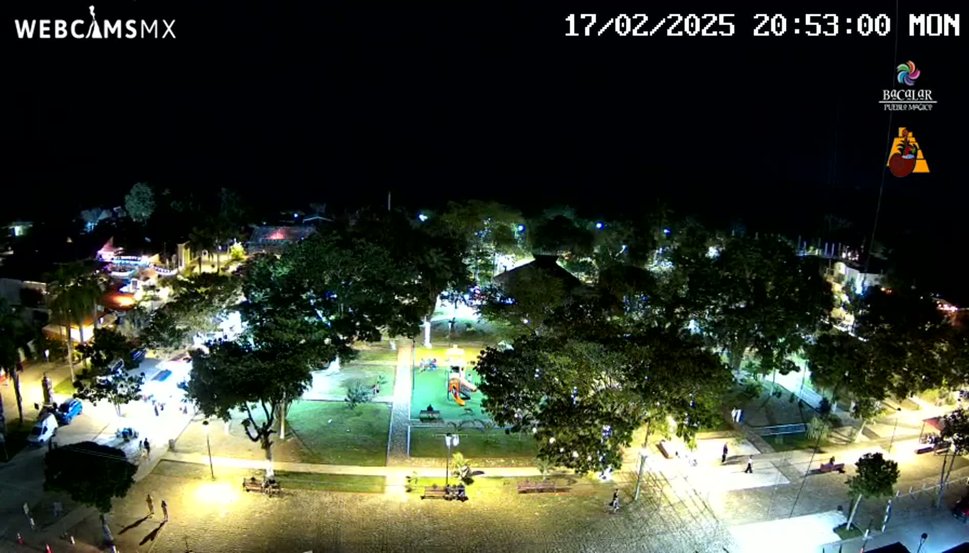 A nighttime aerial view of a town square with a playground, surrounded by illuminated trees and buildings under a clear, dark sky.