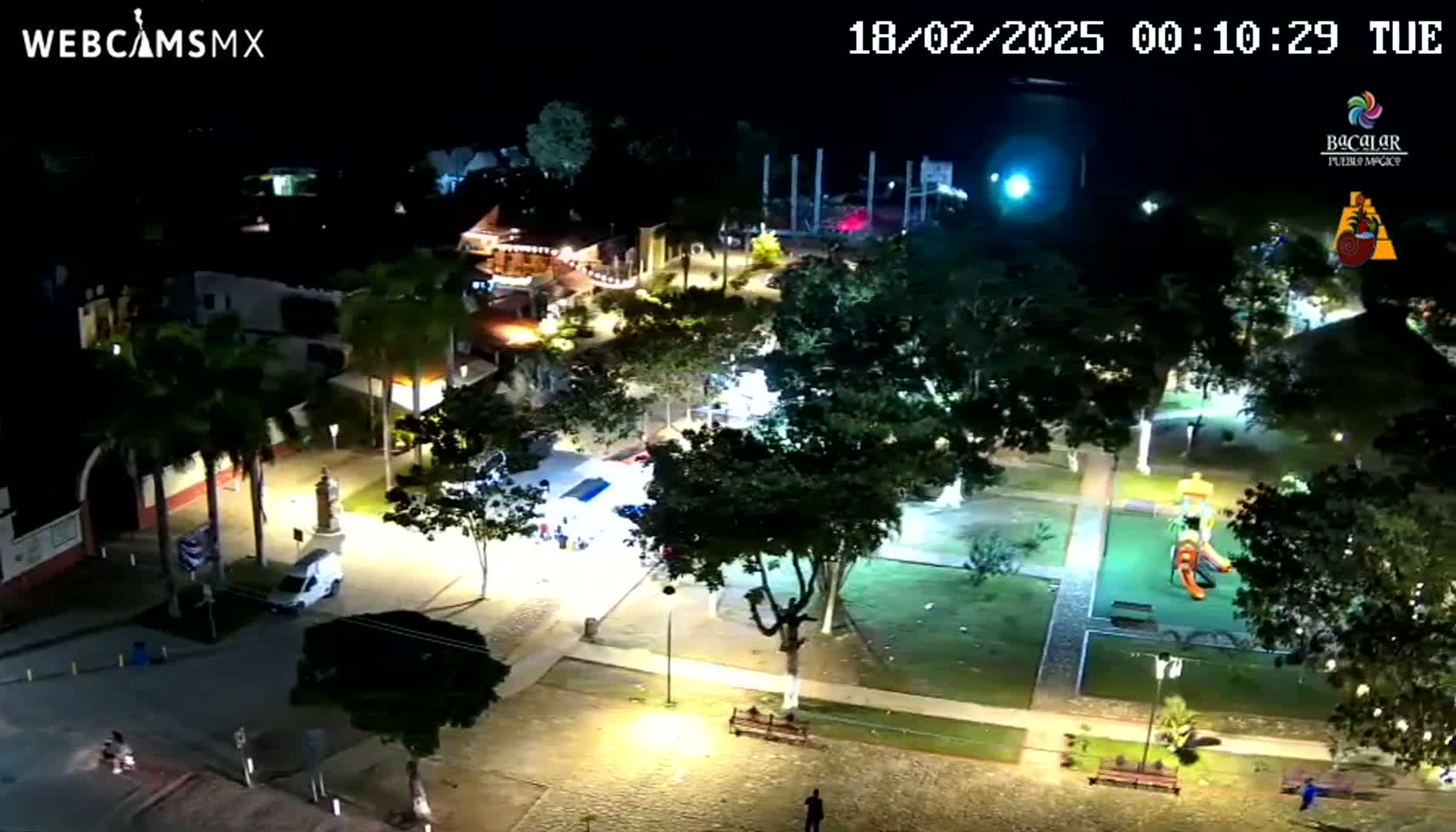 An illuminated town square at night, featuring a park with playground equipment, surrounding buildings, and sparse pedestrian activity under a clear sky.