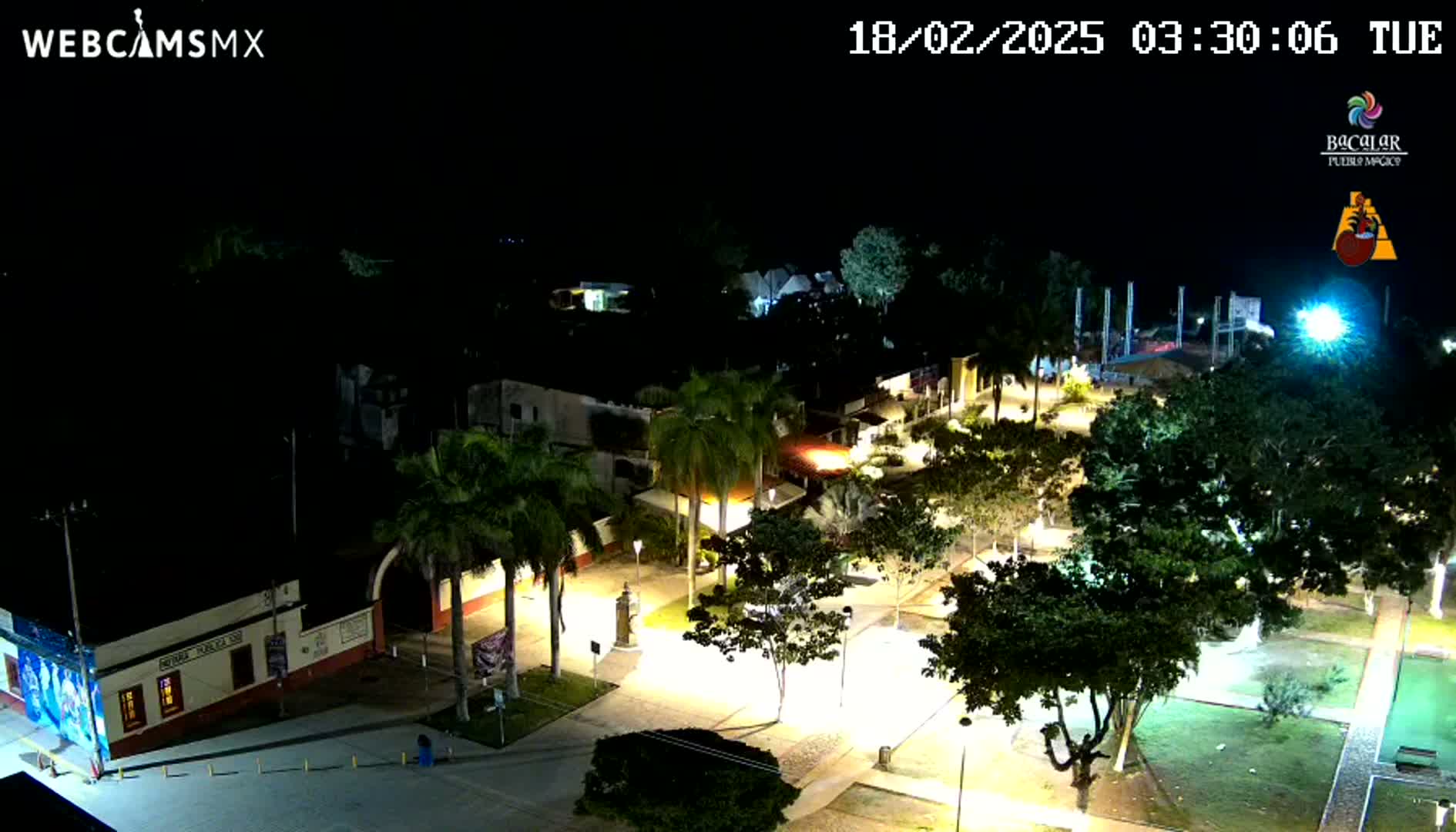 A nighttime, overhead view of a town square illuminated by streetlights, surrounded by palm trees and other lush greenery.