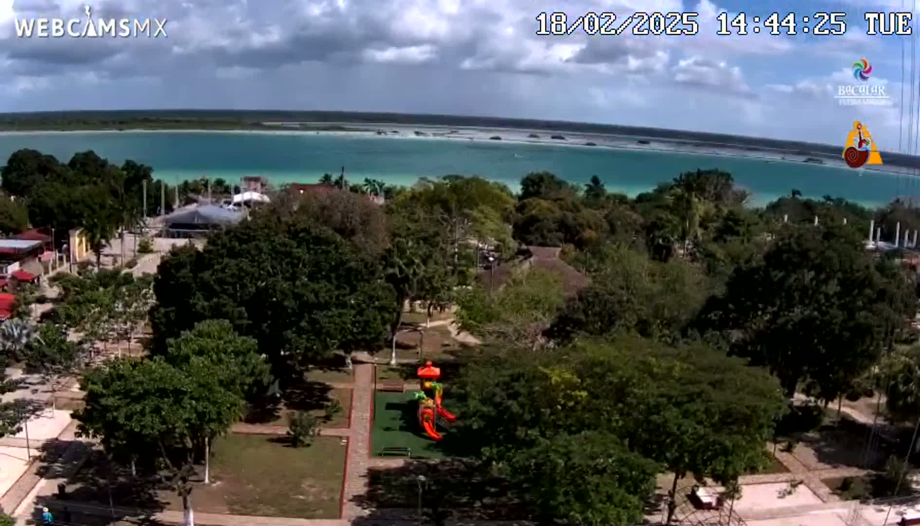 An aerial view shows a town square with a playground next to lush green trees, overlooking a calm, turquoise body of water under a partly cloudy sky.