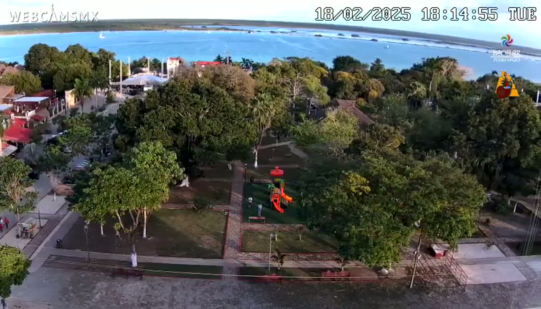 An aerial view shows a town square with a playground and lush green trees, situated beside a body of calm, blue water under a clear sky.