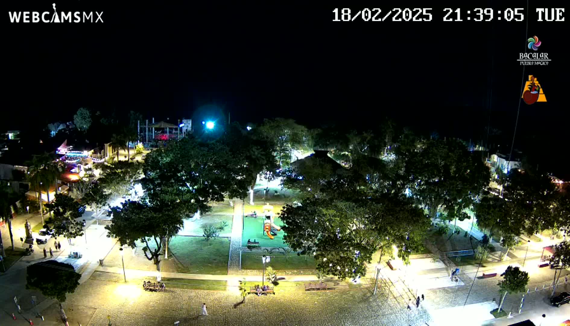 A nighttime aerial view of a town square with a playground, illuminated by streetlights and surrounded by trees.