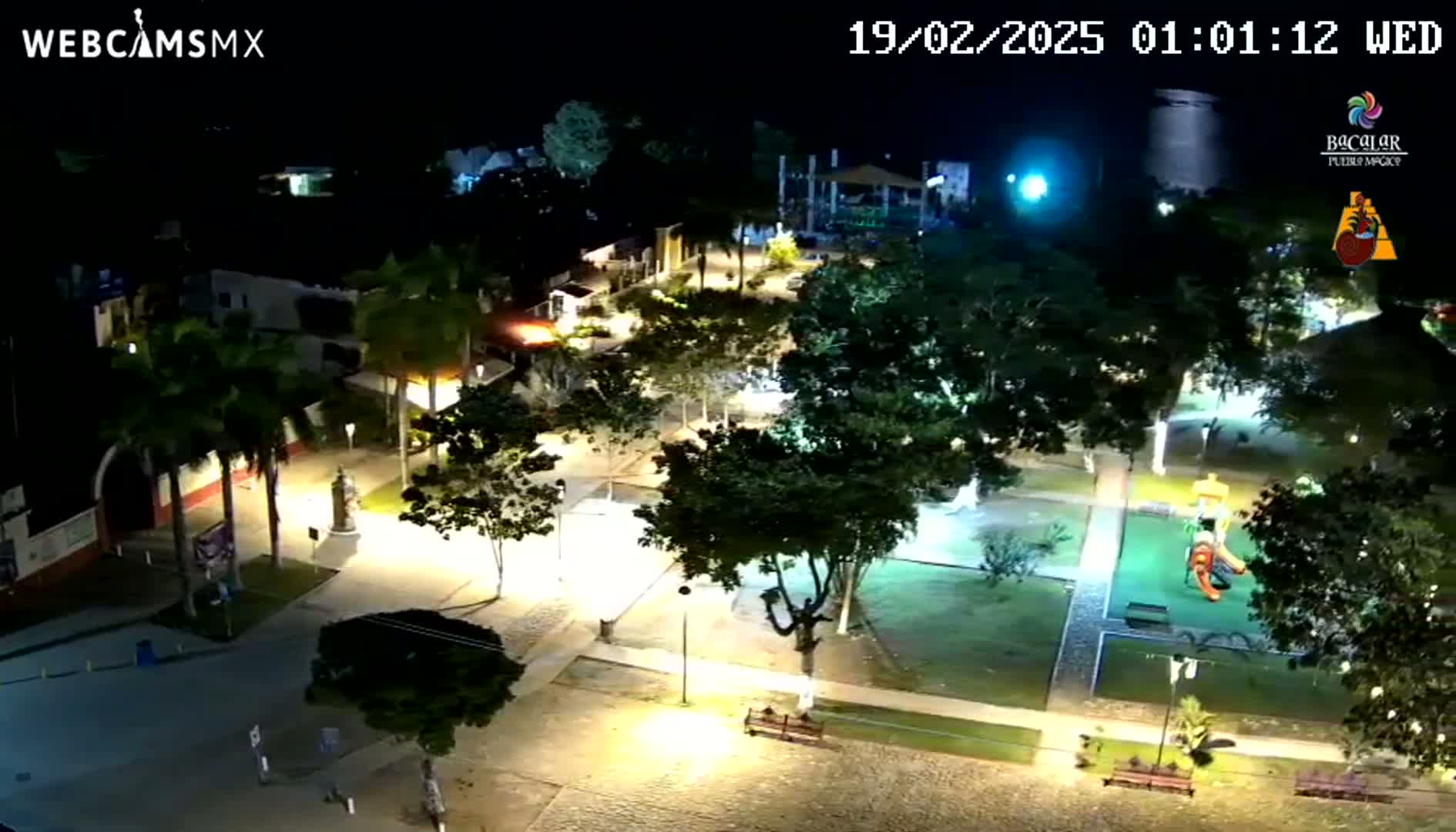 An illuminated town square at night, featuring trees, pathways, and a playground, under clear skies.