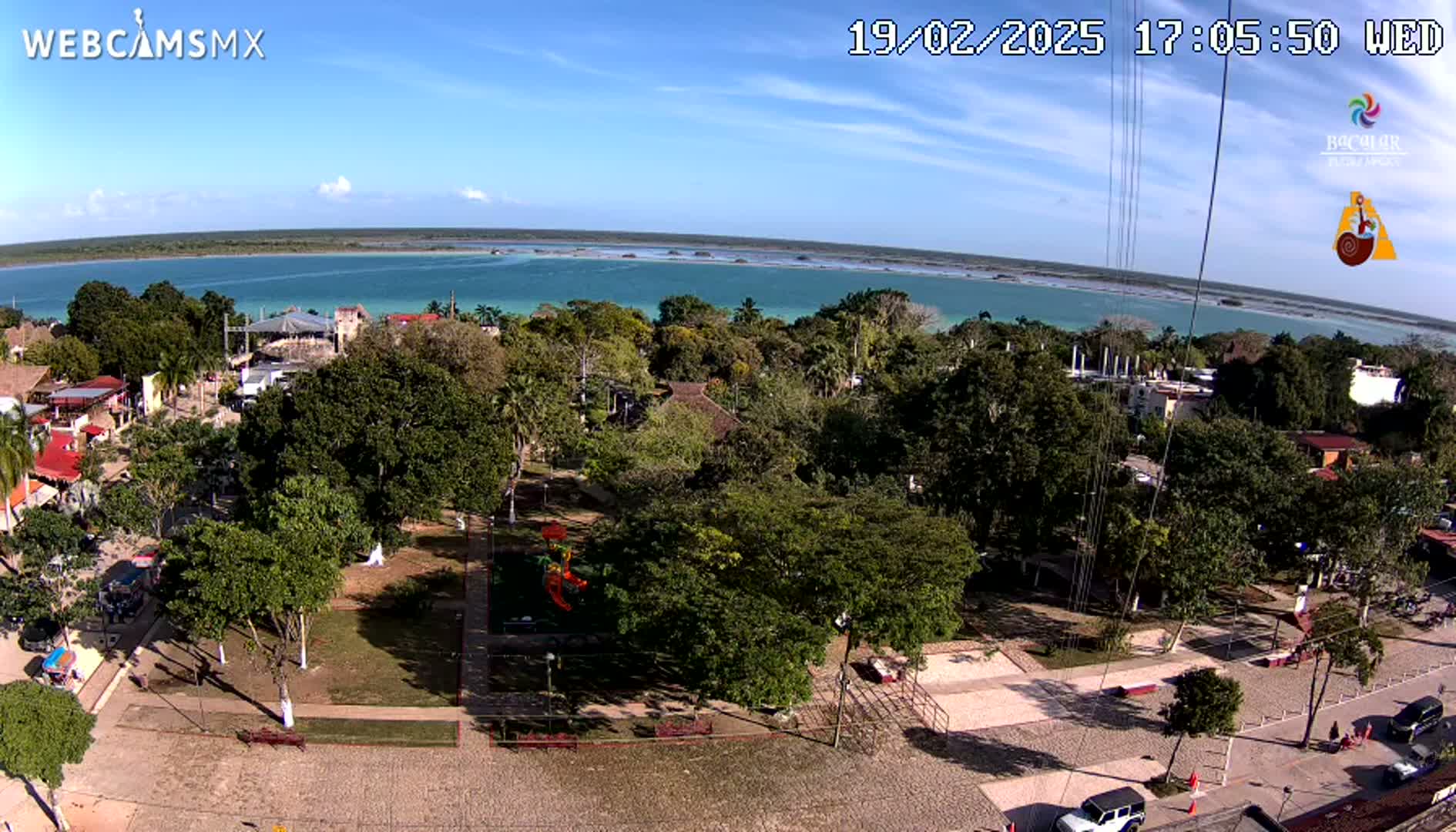 An elevated view shows a town square with lush green trees and a playground, overlooking a calm, turquoise body of water under a sunny sky.