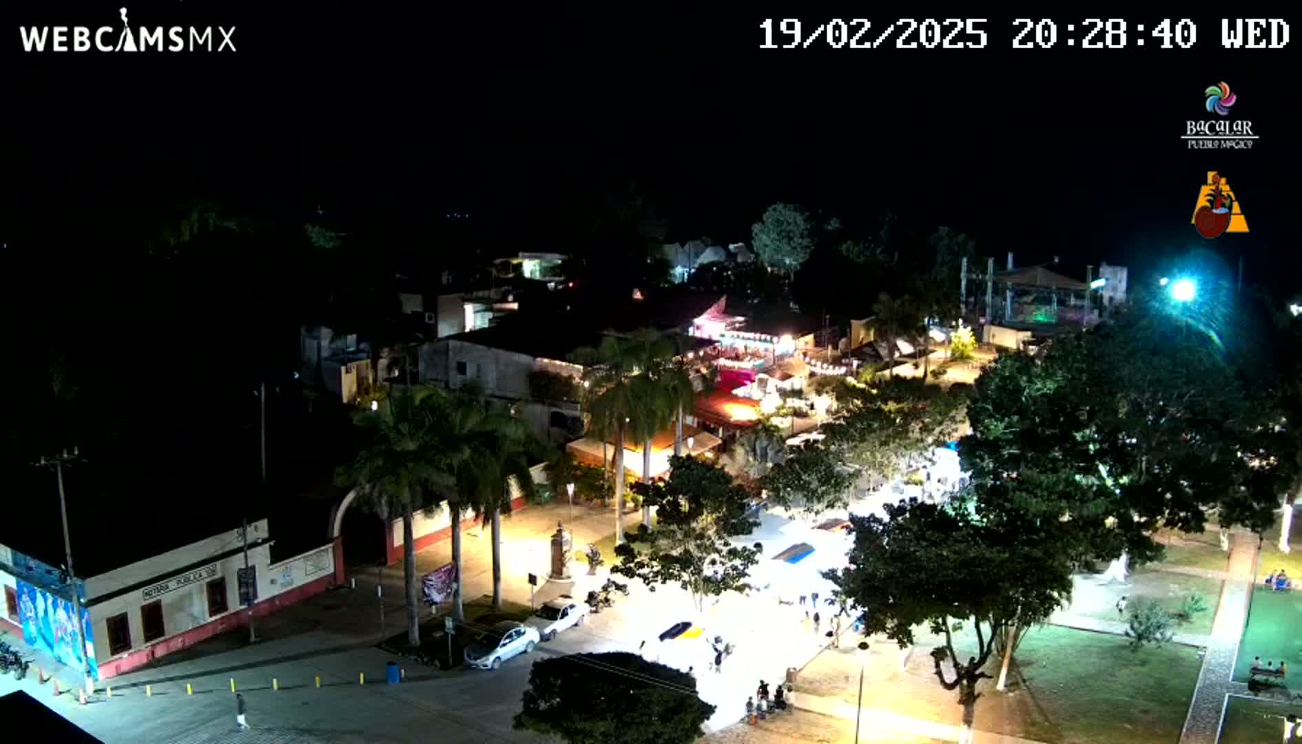 A nighttime aerial view of a town square with illuminated buildings, palm trees, and a few people gathered in the plaza under clear skies.