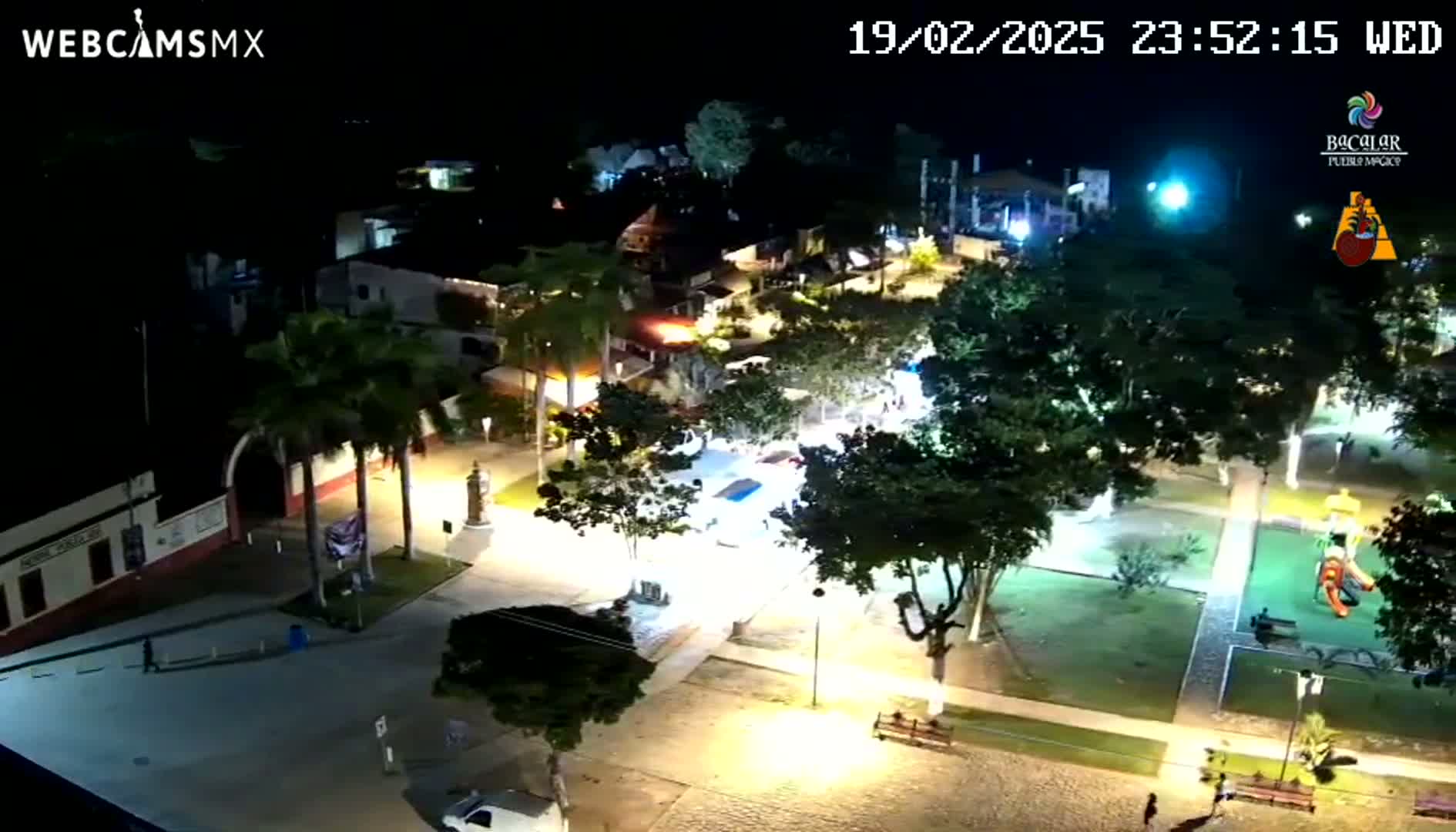 An illuminated town square at night, showing buildings, trees, and a playground, under a clear sky.