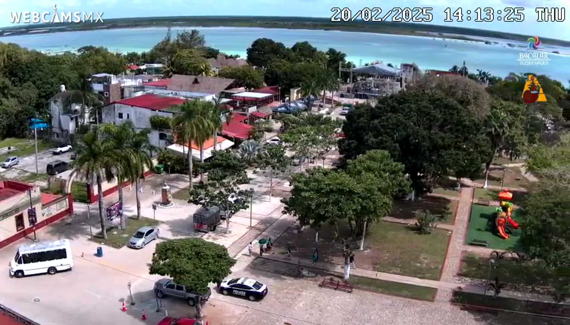 An aerial view of a town square with buildings, parked cars, and a playground, situated next to a body of turquoise water under a mostly clear sky.