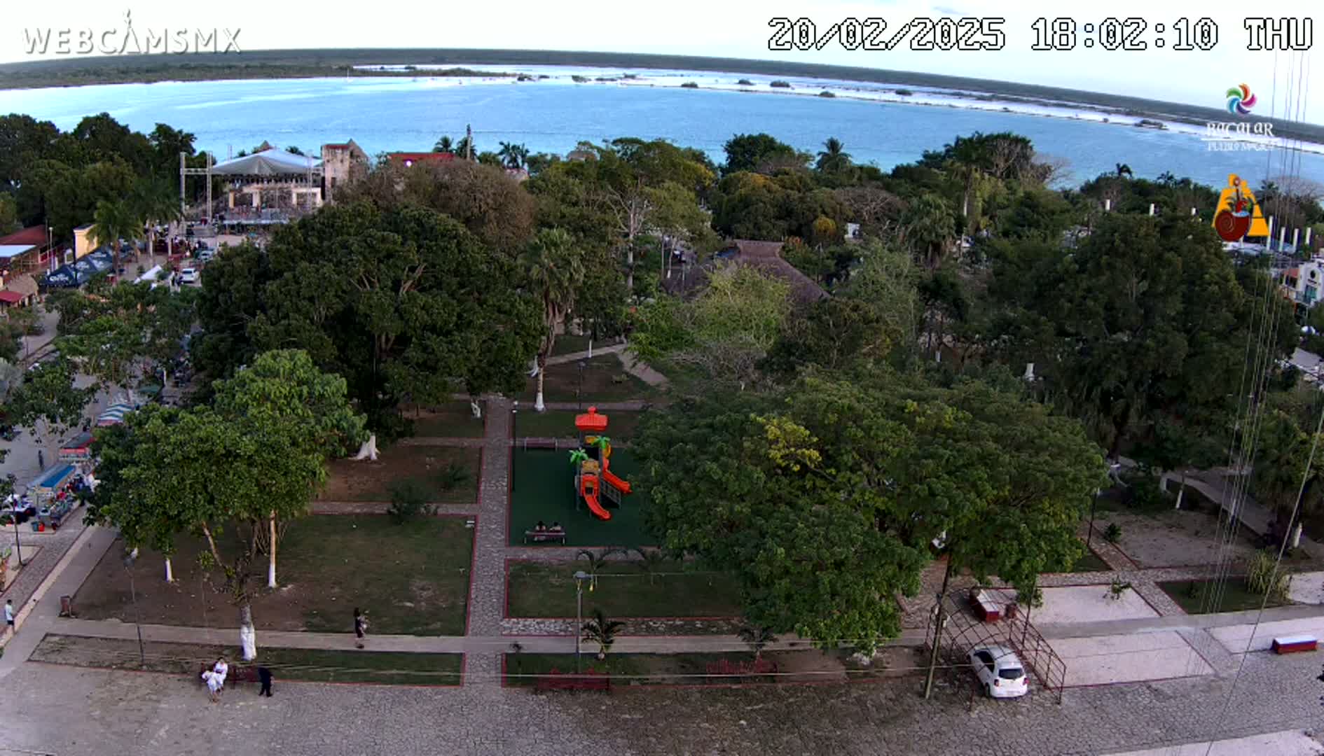 An aerial view shows a park with a playground, surrounded by lush green trees and a paved area, overlooking a calm body of water under a twilight sky.