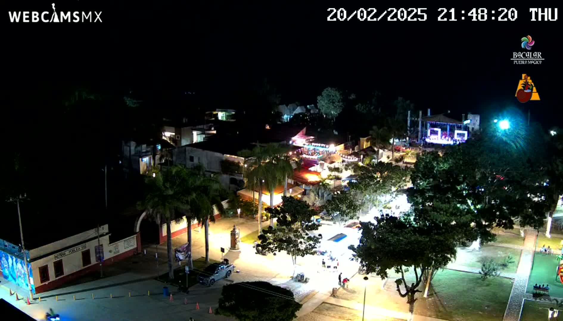 An illuminated town square at night, showing buildings, palm trees, and people gathered under a clear, dark sky.
