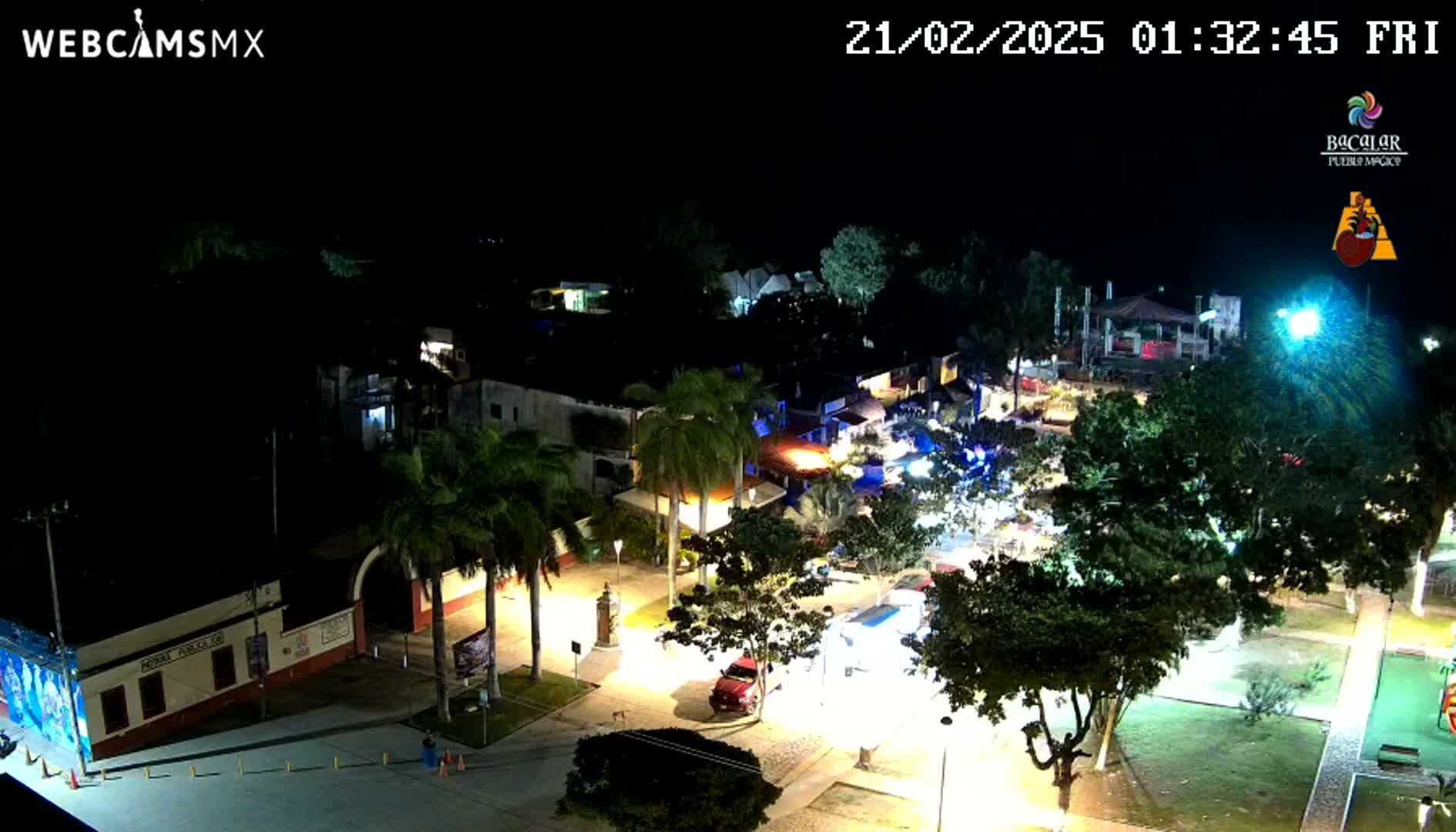 A nighttime, aerial view of a town square with buildings, palm trees, and a few vehicles, illuminated by streetlights.