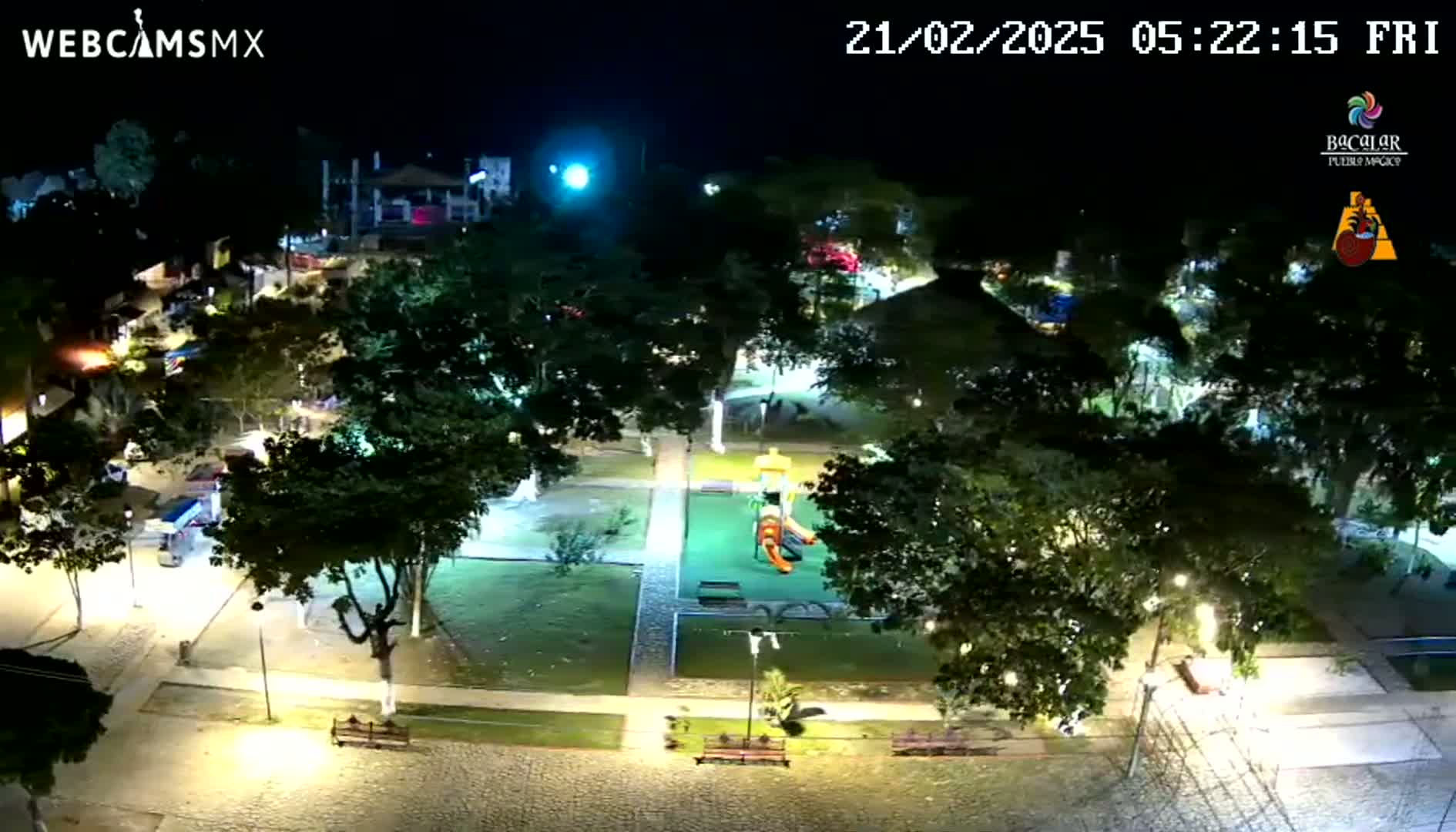 A nighttime aerial view of a park with a playground, illuminated by streetlights, surrounded by trees and paved pathways.