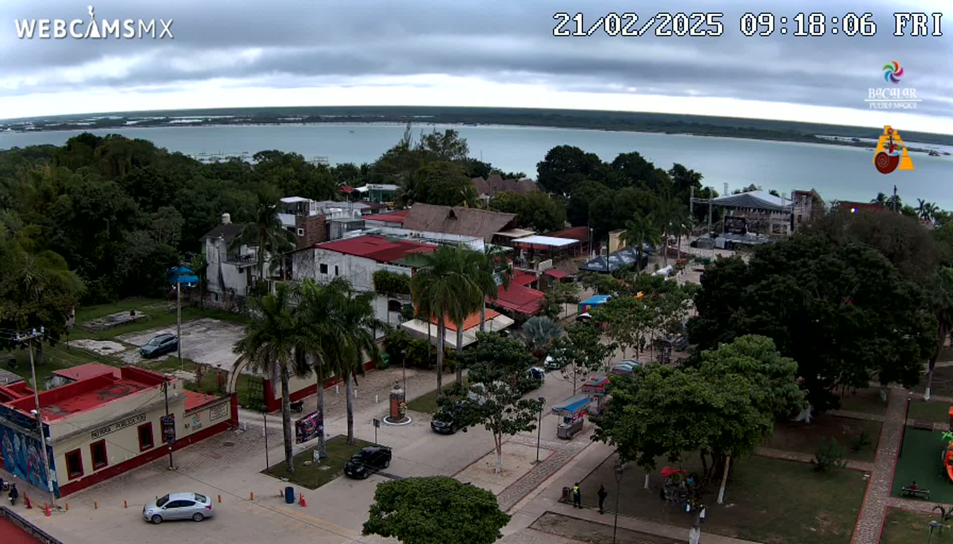 An aerial view shows a town square with palm trees, buildings, and cars, overlooking a calm body of water under a cloudy sky.