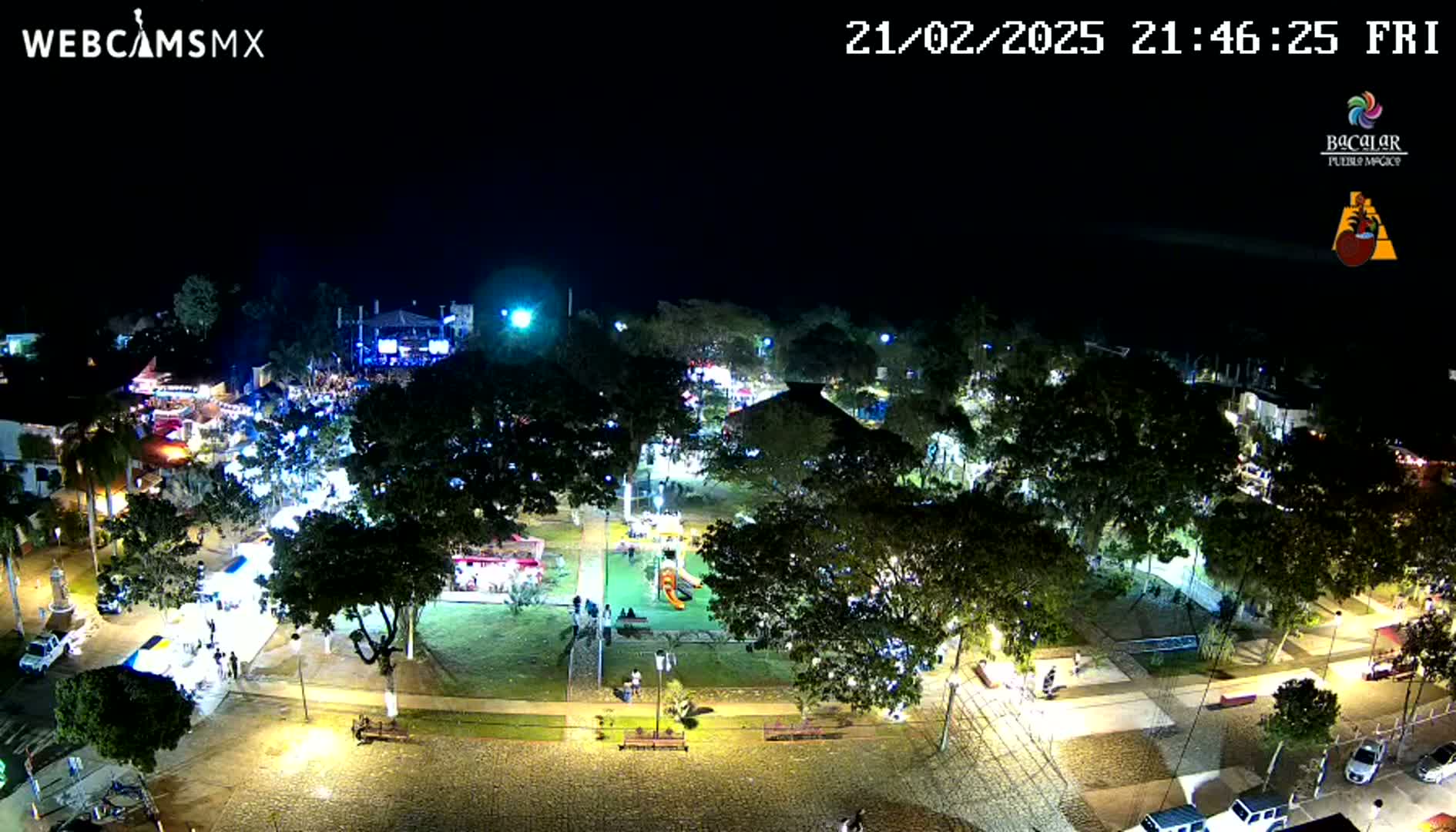 A nighttime aerial view of a town square illuminated by streetlights, featuring a park with trees and some people, surrounded by buildings and roads.